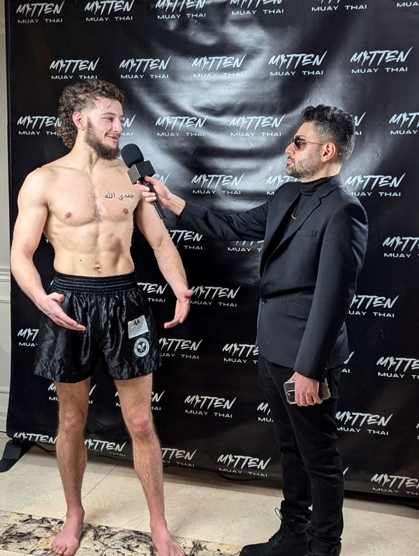 Coach Elijah Halloun being interviewed at a Mitten Muay Thai event, wearing Fight Mechanix shorts