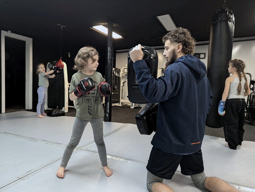 A young student in boxing gloves faces a coach who kneels to hold pads at her height